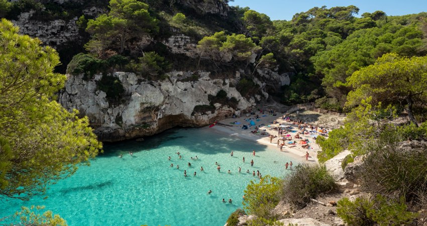 Aerial view of Cala Macarelleta, with turquoise waters and families enjoying a day at the beach among the cliffs.