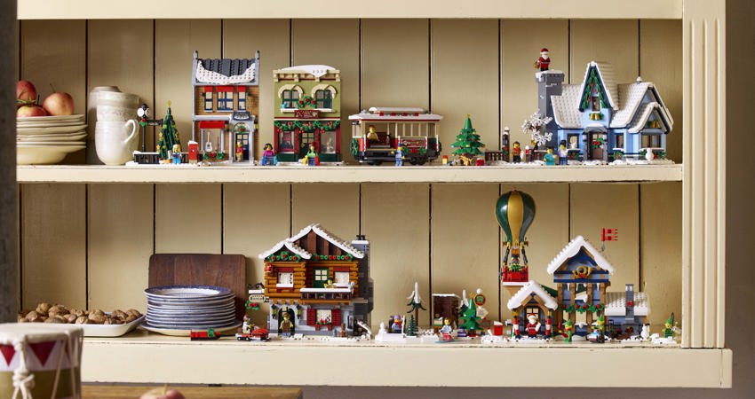Consigue hasta 10 Avios por cada 1€ que gastes con Lego. En la imagen, las construcciones de Lego de la colección de Navidad sobre una estantería de madera, rodeadas por platos y vasos de temática navideña, con manzanas y galletas recién horneadas 