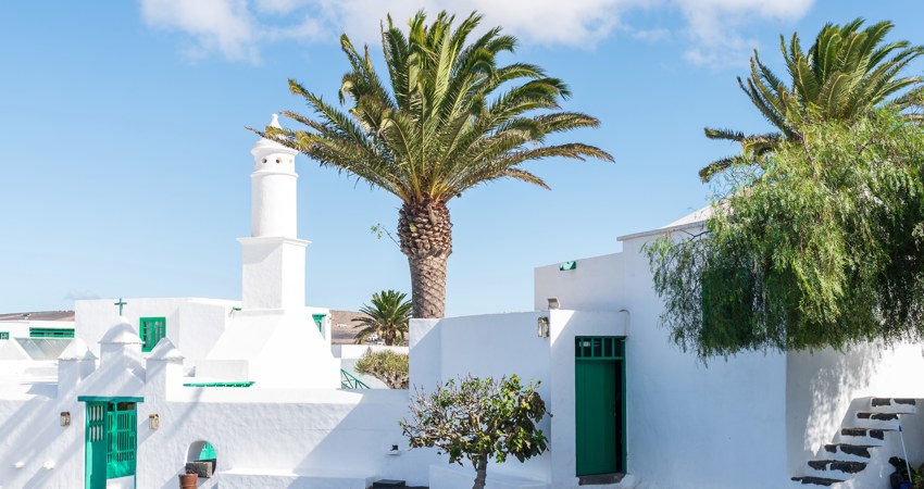 En la imagen, una de las plazas de San Bartolomé, con bajas casitas bajas salpicadas de palmeras. En el fondo, el perfil del Monumento a la Fertilidad de César Manrique