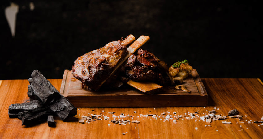 Presentación de un costillar a la brasa en La Carnicería, sobre una tabla de madera, acompañado de un bodegón de sal y carbón