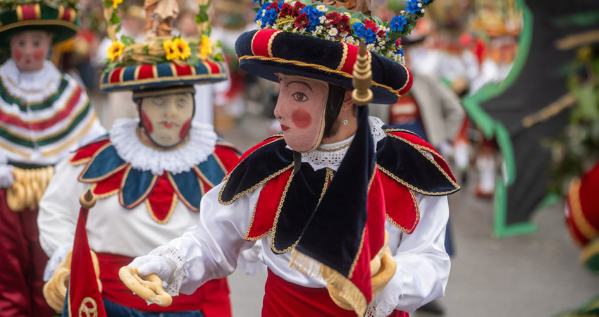 En primer plano, un participante de los carnavales porta una gran máscara tradicional, junto a sombrero de colores azul y rojo y camisa blanca