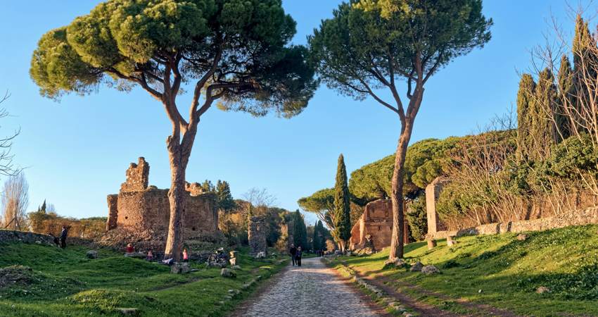 Vista de la calzada de teselas de la Via Appia, rodeada de naturaleza, con árboles y restos arqueológicos a sendos lados de la vía