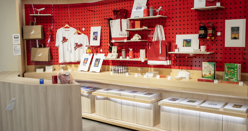 Espacio Iberia Madrid 2025. En la imagen, la tienda del Espacio Iberia, con camisetas, láminas y bolsos con el estilo corporativo de Iberia, incluyendo una réplica del bolso que portan los tripulantes a bordo