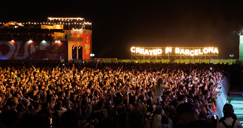 Primavera Sound 2025. In the image, dark night with thousands of people dancing and singing in front of a stage. In the background, a sign with yellow neon lights where you that reads “Created in Barcelona”.