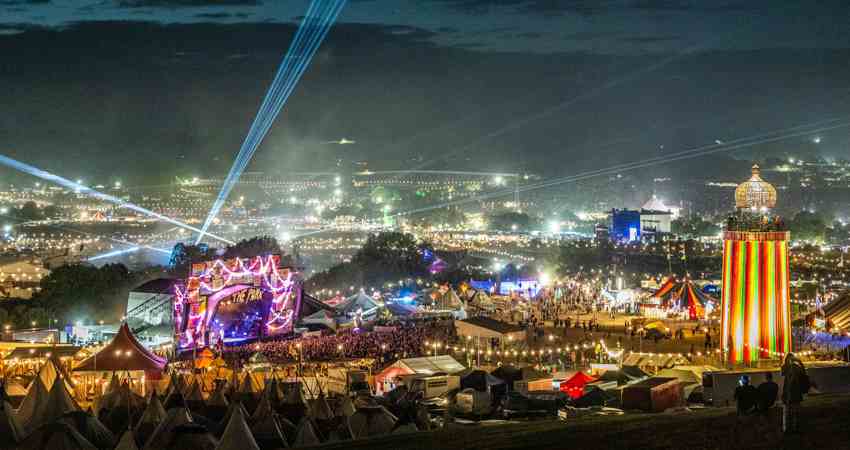 En la imagen, un mar de luces con un faro de colores y un escenario masivo con luces de neón, todo rodeado por carpas y tiendas de campaña.