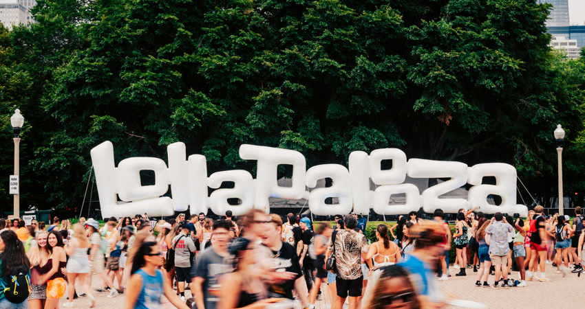 En la imagen, letras gigantes blancas donde se lee Lollapalooza. Vegetación al fondo y visitantes alrededor.