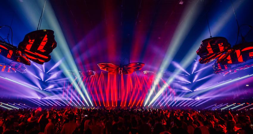 The image shows giant butterflies flying over the crowd in front of the stage, pursued by columns of light.
