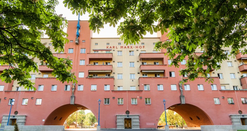 En la imagen, una de las entradas de Karl-Marx-Hof, con los árboles que rodean el edificio. Pueden verse las arcadas de acceso a los parques interiores. Entre ellas, los diferentes portales a las viviendas, en edificios blancos y rojos.