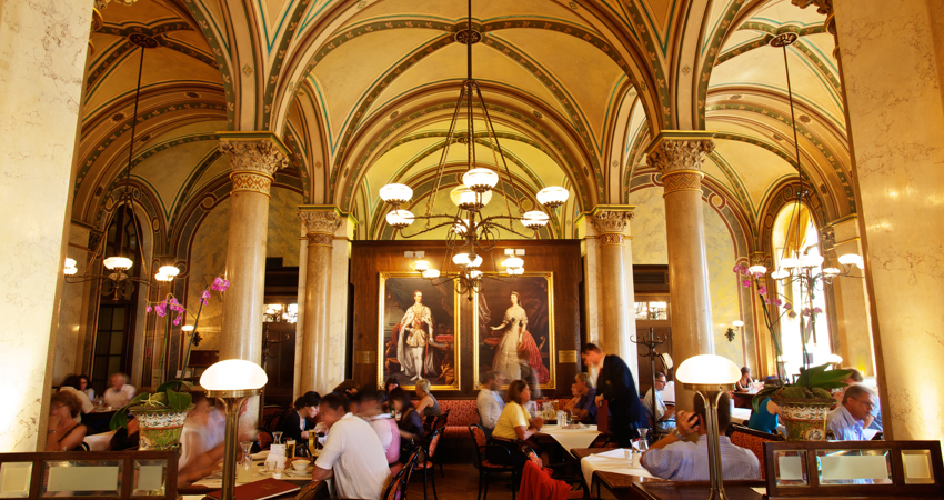 Café Central en Viena. Al fondo, dos retratos de Francisco José I de Austria y la emperatriz Elisabeth (Sissi). Mesas llenas de clientes disfrutan del ambiente elegante bajo lámparas colgantes clásicas.