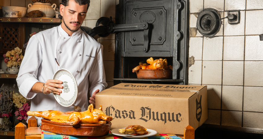 En la imagen, un cocinero de Casa Duque se dispone a partir un cochinillo recién asado sobre bandeja de barro a la manera tradicional (con un plato). Al lado, una de las cajas de comida a domicilio de Casa Duque y, tras ella, un horno de hierro donde se cocina otra pieza
