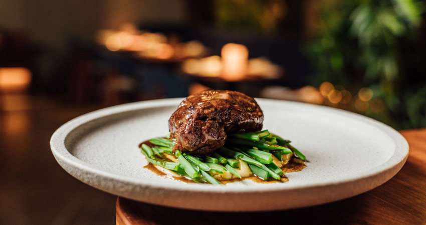 In the image, pork cheeks on a bed of vegetables in a grey ceramic dish, against a background decorated with Christmas candles