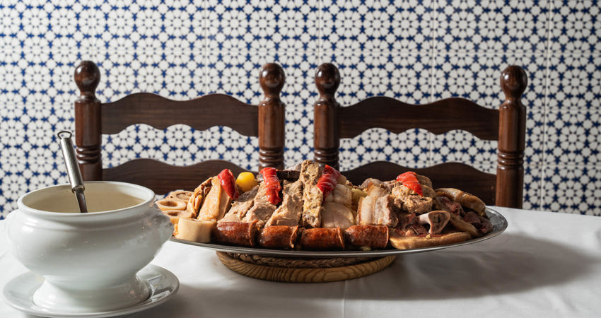 In the foreground, a table with a white tablecloth, a soup tureen and a large platter of meat. In front of them are two wooden and wicker tavern chairs and a background of blue and white tiles with geometric patterns