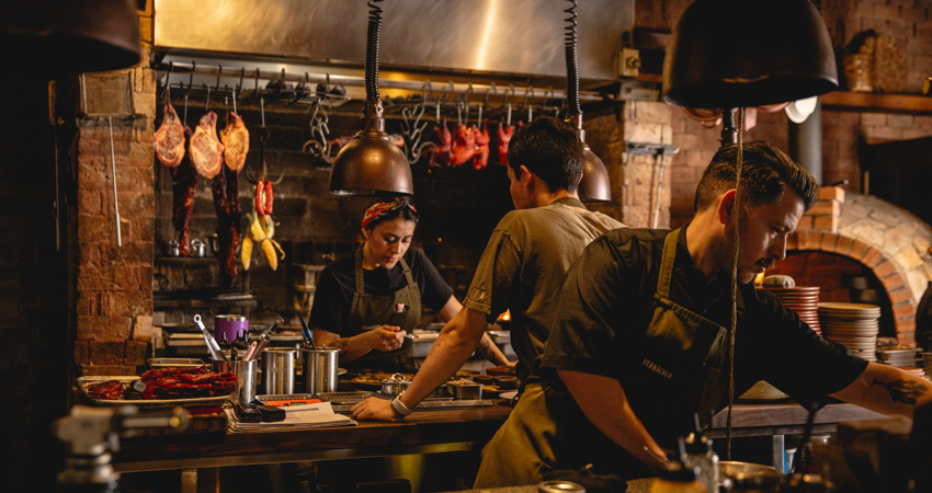 Vista del área de preparación en Vernáculo, con su estilo industrial. En primer plano, un chef de perfil organiza la mesa; al fondo, una mujer con pañuelo en la cabeza y otro cocinero trabajan bajo lámparas cálidas, rodeados de cortes de carne y pollos colgados sobre una parrilla de ladrillo.