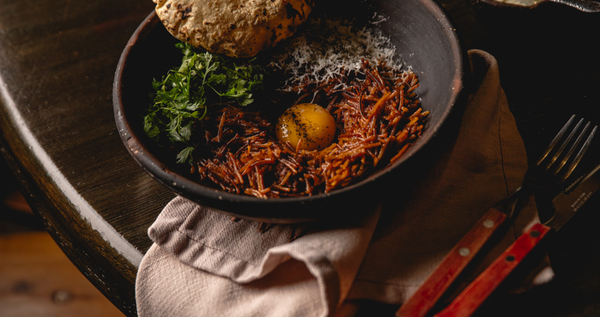 Fotografía cenital de un cuenco de barro oscuro sobre una mesa de madera. El plato contiene fideos rojos tipo "fideo seco", coronados con una yema de huevo cruda, queso rallado, cilantro fresco y una tortilla de maíz tostada al fuego.